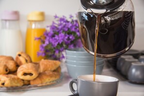 A cup of coffee being poured from a metallic pot, accompanied by a plate of pastries and a bottle of orange juice.