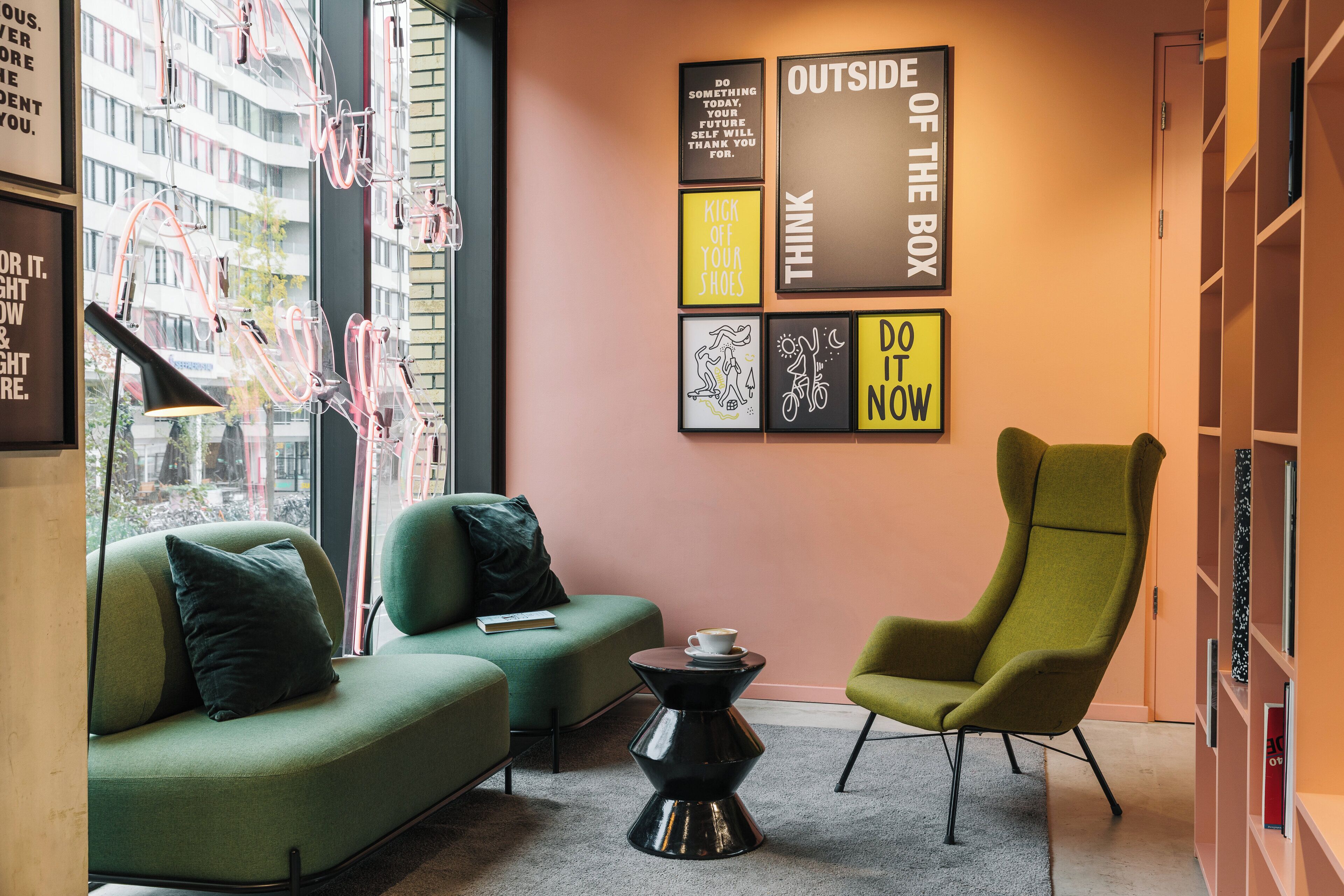 Un espace de détente moderne avec un canapé vert, un fauteuil vert et une table basse noire. Des affiches motivantes sont accrochées au mur, et une fenêtre offre une vue sur les bâtiments.