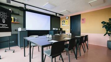 Un salón de conferencias con una larga mesa, sillas, una pantalla de proyector, un reloj y una planta.