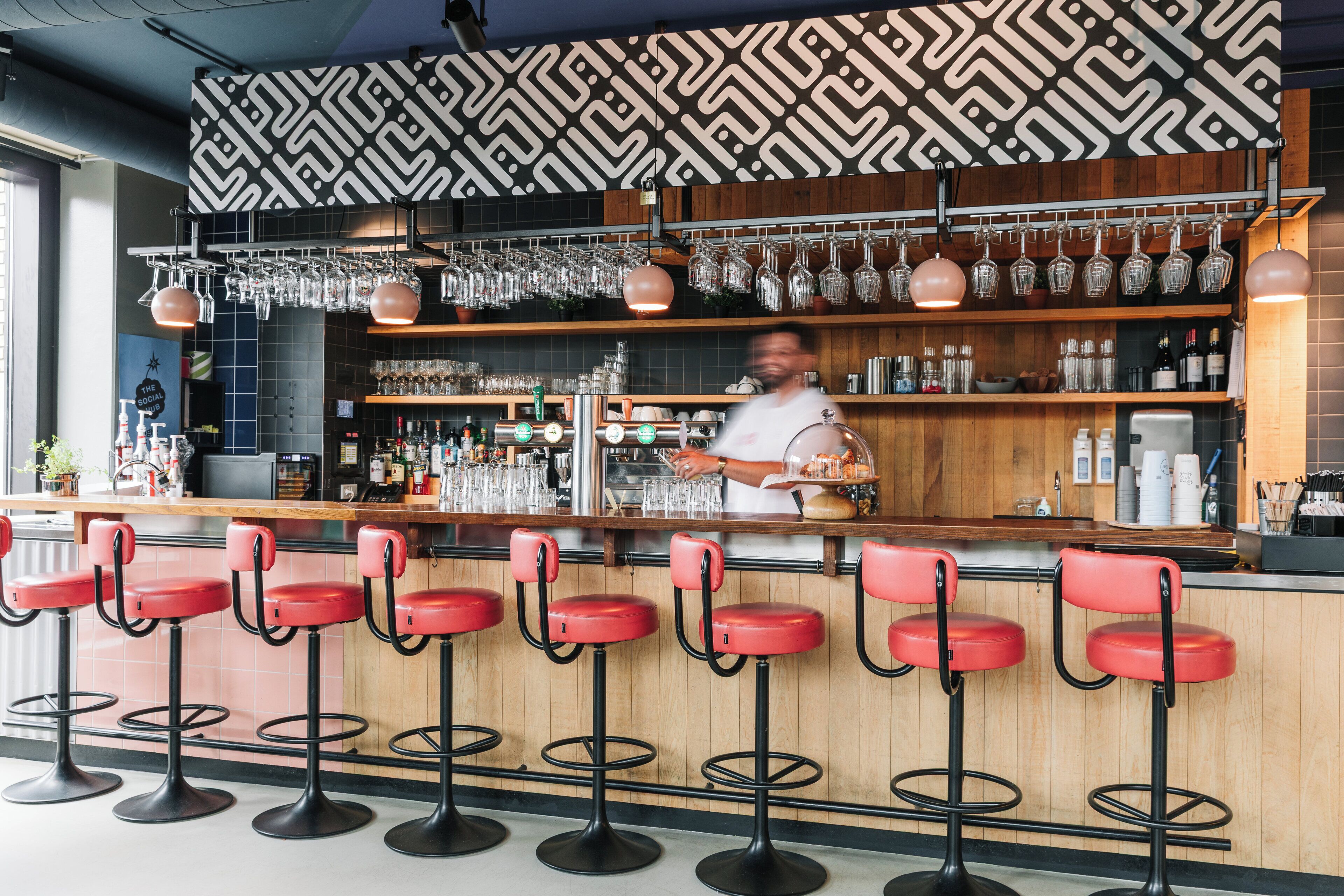 Zona de bar con varios taburetes, un cantinero y estanterías llenas de botellas y vasos.