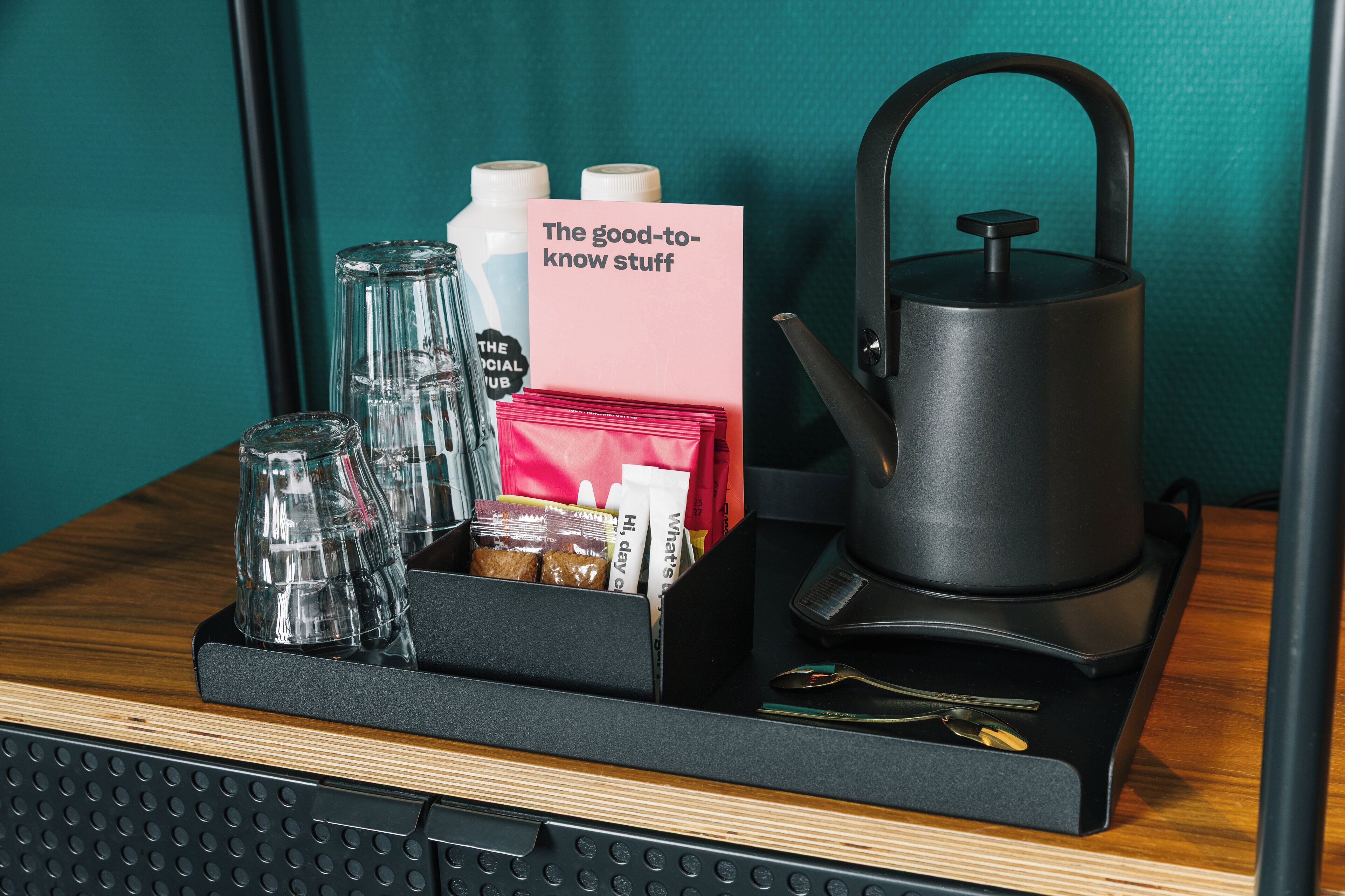 A tray with a kettle, a glass, a bottle, a card, and some snacks on a wooden surface.