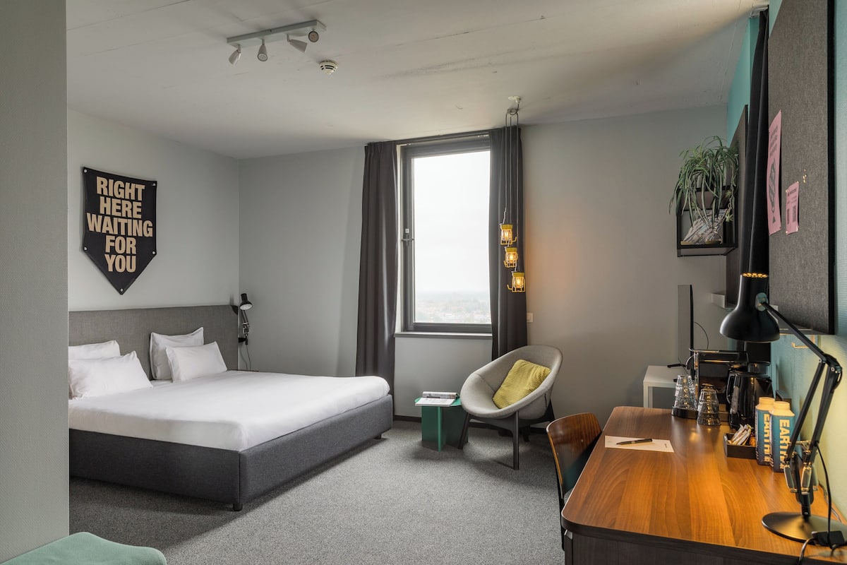 Ett modernt hotellrum med en säng, ett skrivbord, en stol, ett litet bord och ett fönster med gardiner.