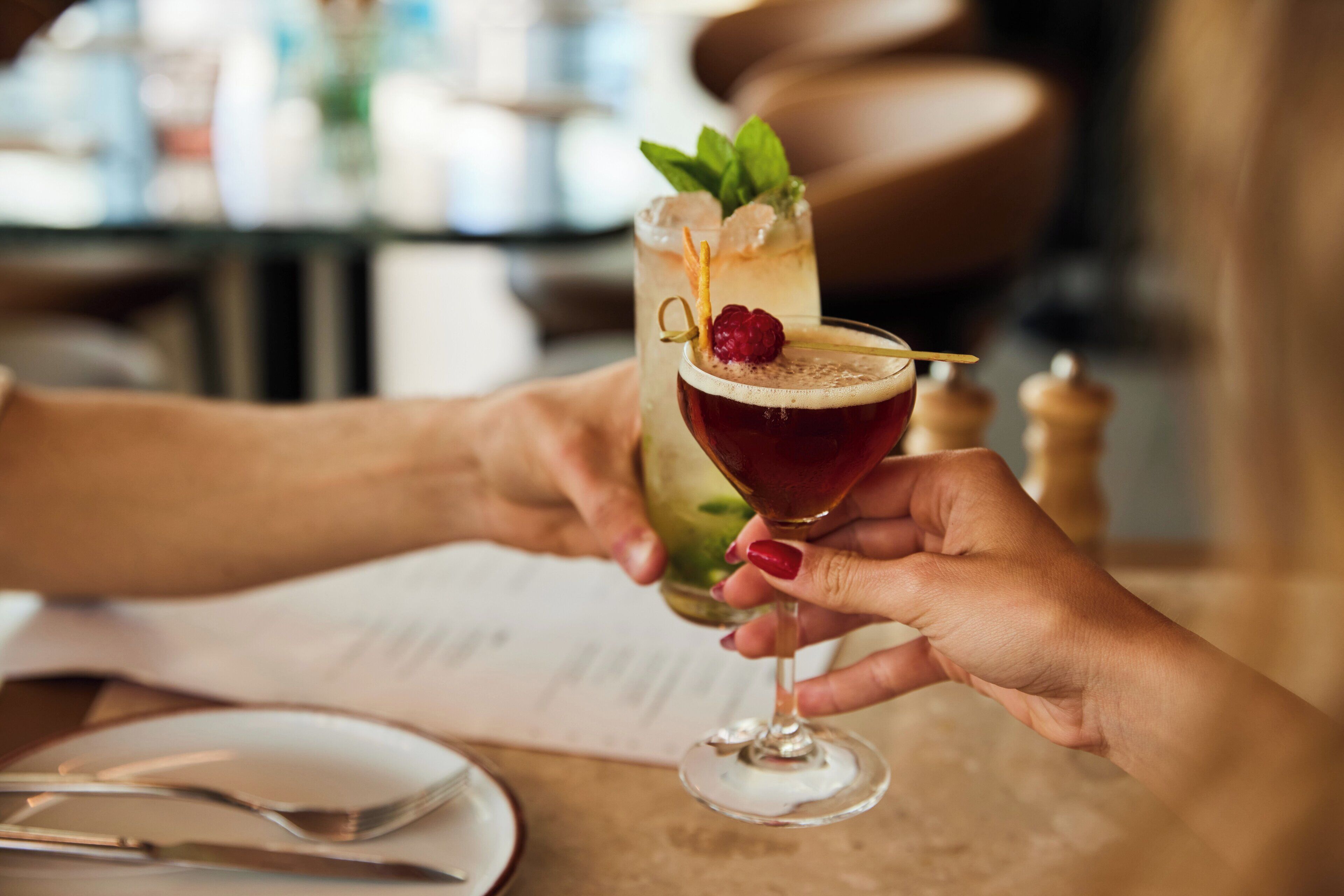 A person holding a cocktail glass with a garnish, another person holding a tall glass with a drink, and a menu on the table.
