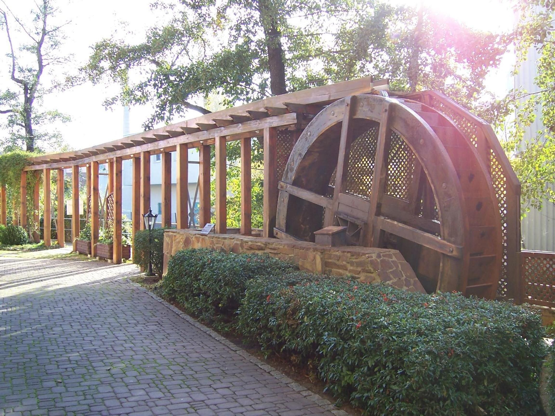 Eine Holzhütte mit einem großen Wasserrad, umgeben von Steinmauern und Grünpflanzen.