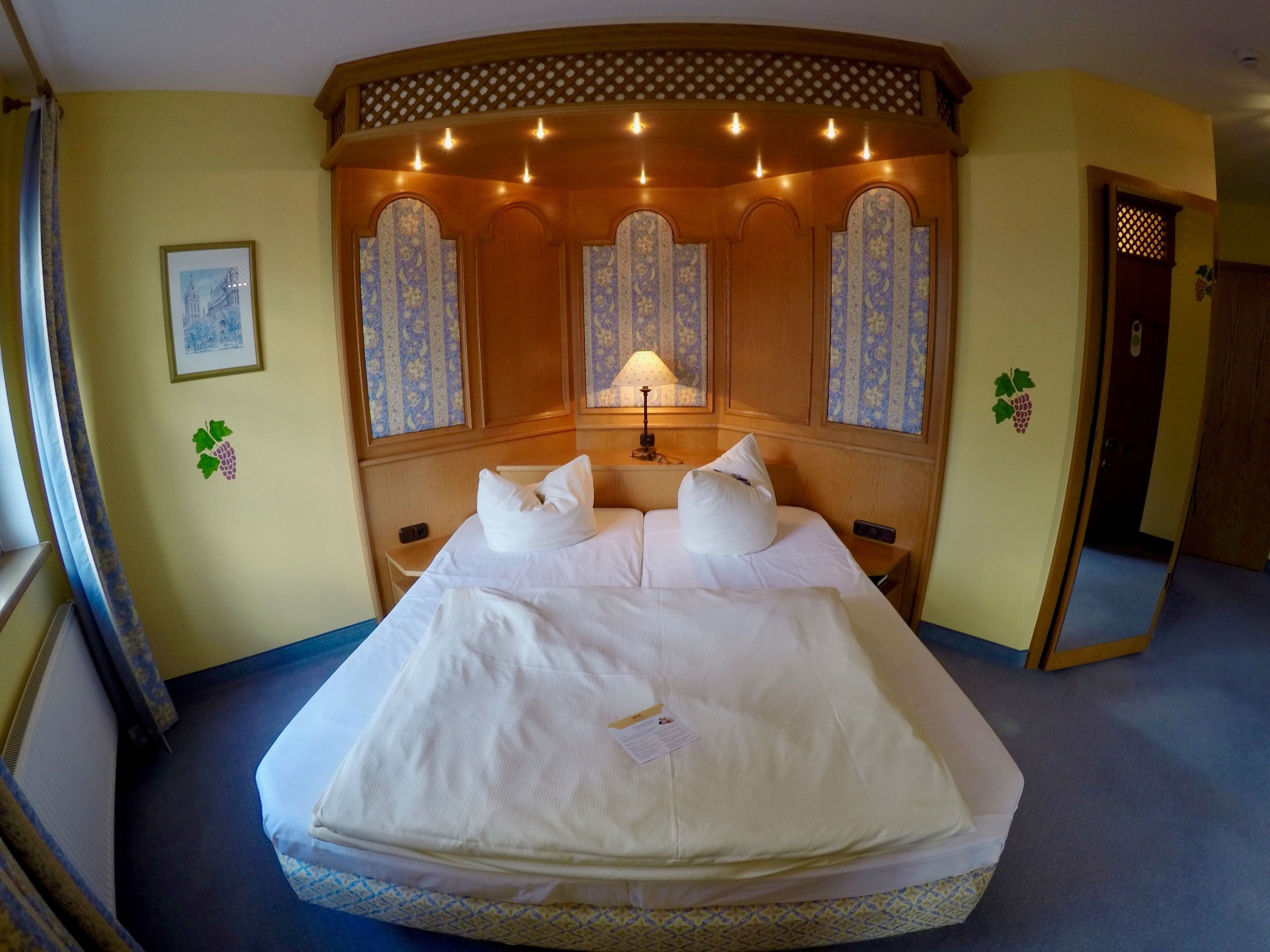 A hotel room with a large bed, a headboard with decorative panels, a bedside table with a lamp, and a framed picture on the wall.