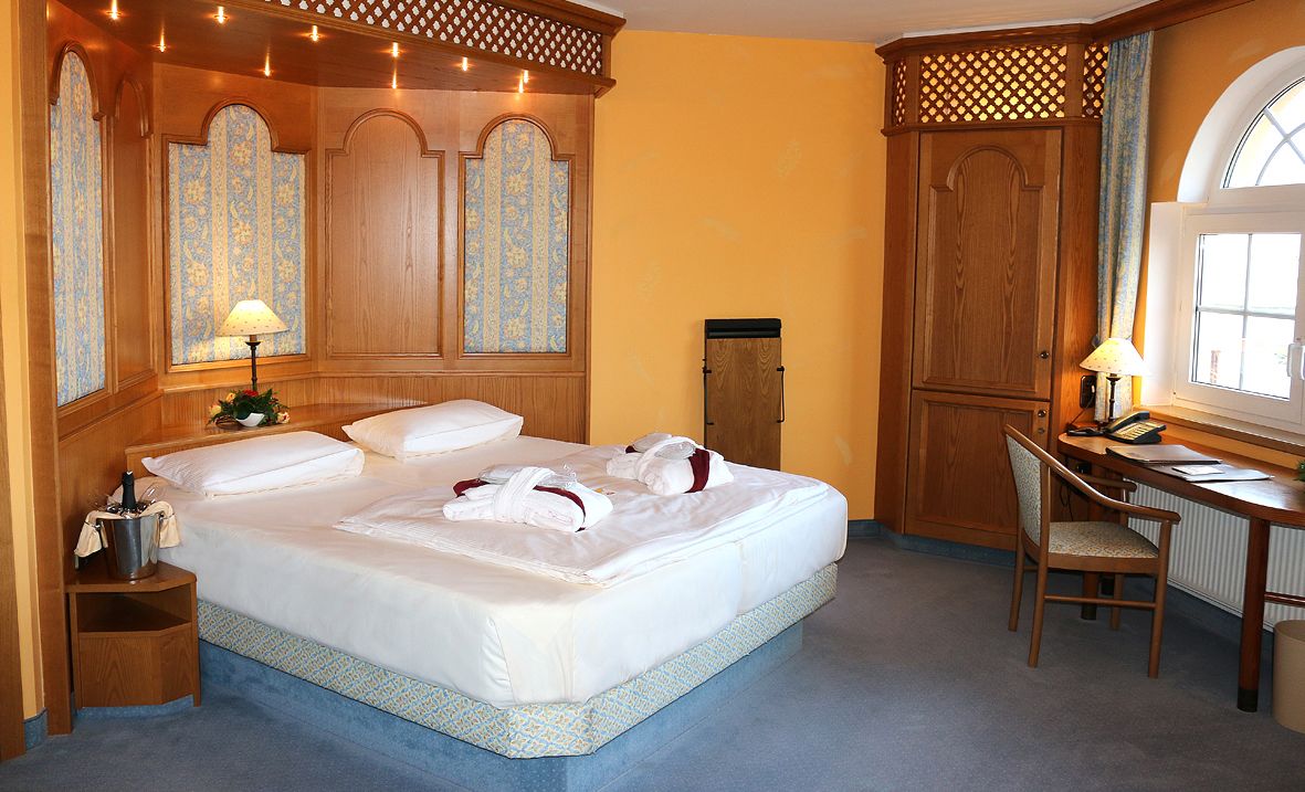 A hotel room with a large bed, wooden headboard, a desk with a chair, and a window with curtains.