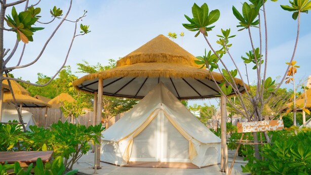 Une tente à toit de chaume, dotée d’un auvent en toile, entourée d’une végétation luxuriante et agrémentée d’un panneau en bois indiquant « Camp de plage de Bangsalen ».