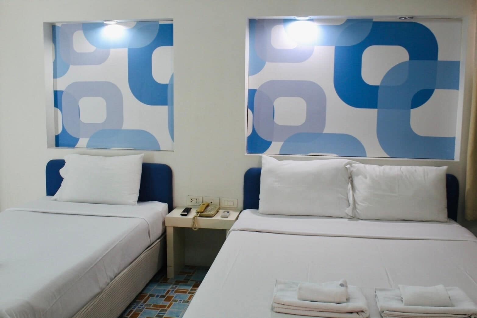 A hotel room with two beds, each with white bedding and pillows. There is a nightstand between the beds with a telephone and a lamp. The walls have a blue abstract pattern.