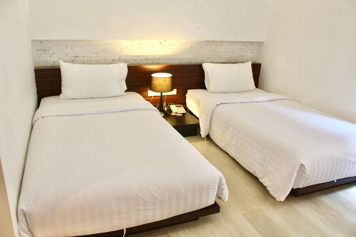 A hotel room with two single beds, a nightstand with a lamp, and a white brick accent wall.