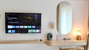 Une chambre d’hôtel moderne équipée d’un téléviseur à écran plat diffusant un film, d’un miroir, d’une lampe de chevet et d’une petite table sur laquelle se trouvent un vase et des télécommandes.