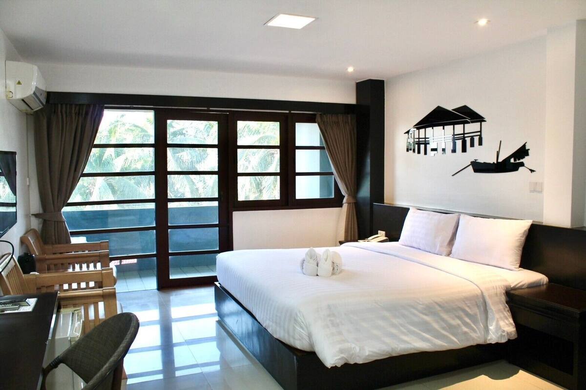 A hotel room with a bed, a desk with a chair, a large window with curtains, and a wall-mounted artwork of a boat.