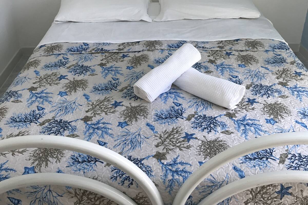 Ein Bett mit blumigem Überwurf, zwei Kissen und einem zusammengerollten Handtuch. Vor dem Bett stehen zwei weiße Metallstühle.