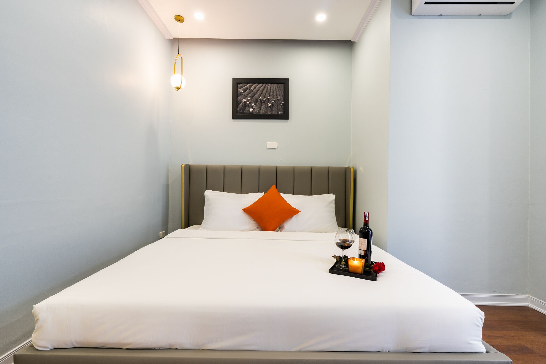 A neatly made bed with white bedding and an orange pillow, a bedside table with a wine bottle and candle, and a framed picture on the wall.