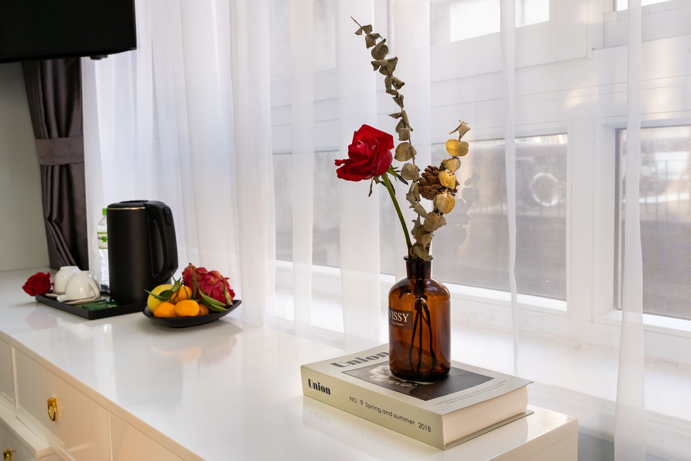 Une table blanche sur laquelle se trouve un livre intitulé « Union, numéro 9, printemps-été 2018 », un vase marron contenant des fleurs séchées et une machine à café.