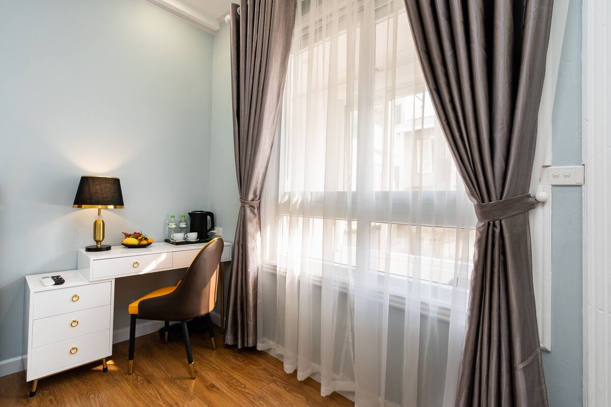 A hotel room with a desk, chair, and a lamp. There is a window with curtains.