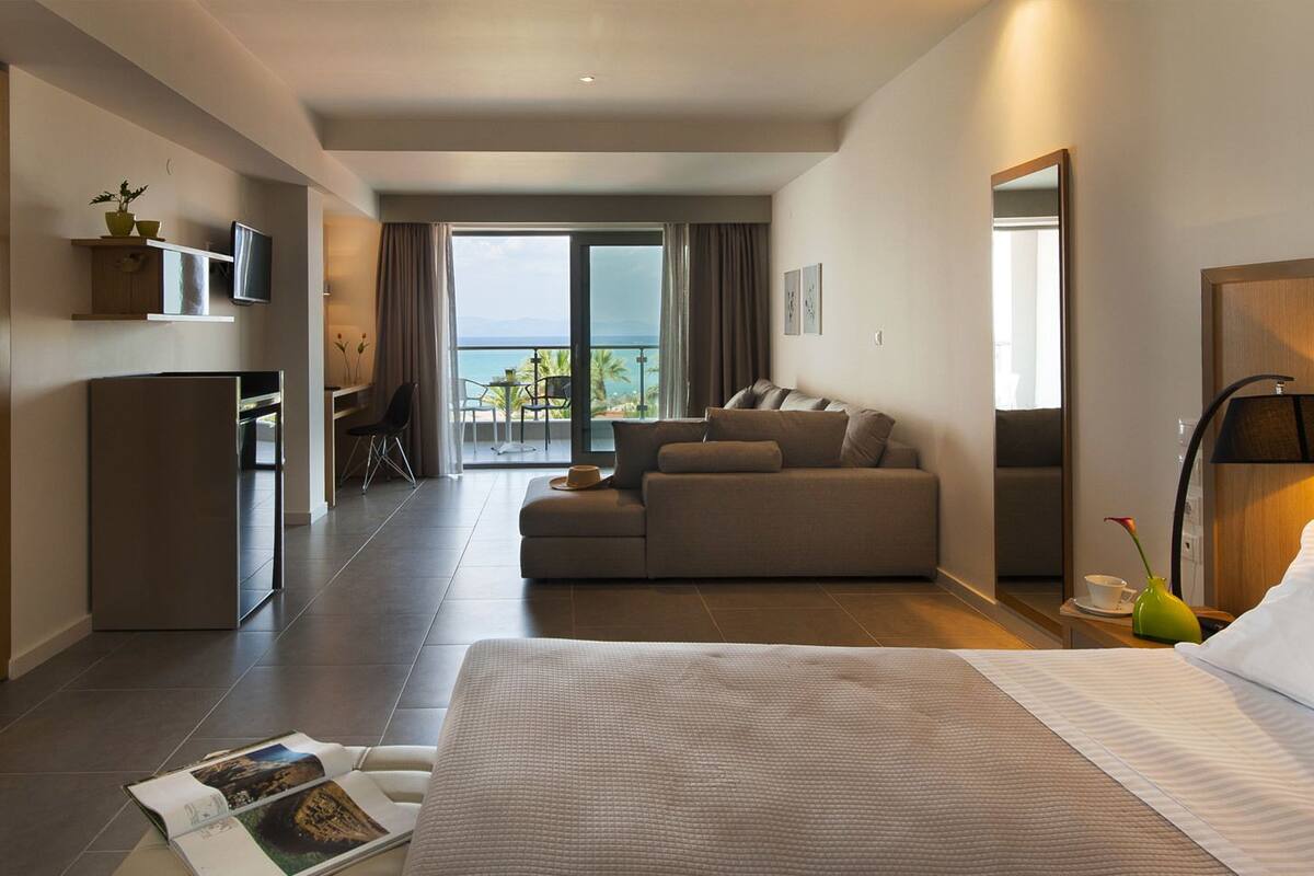 A modern hotel room with a large bed, a sofa, a desk, and a balcony with a view.