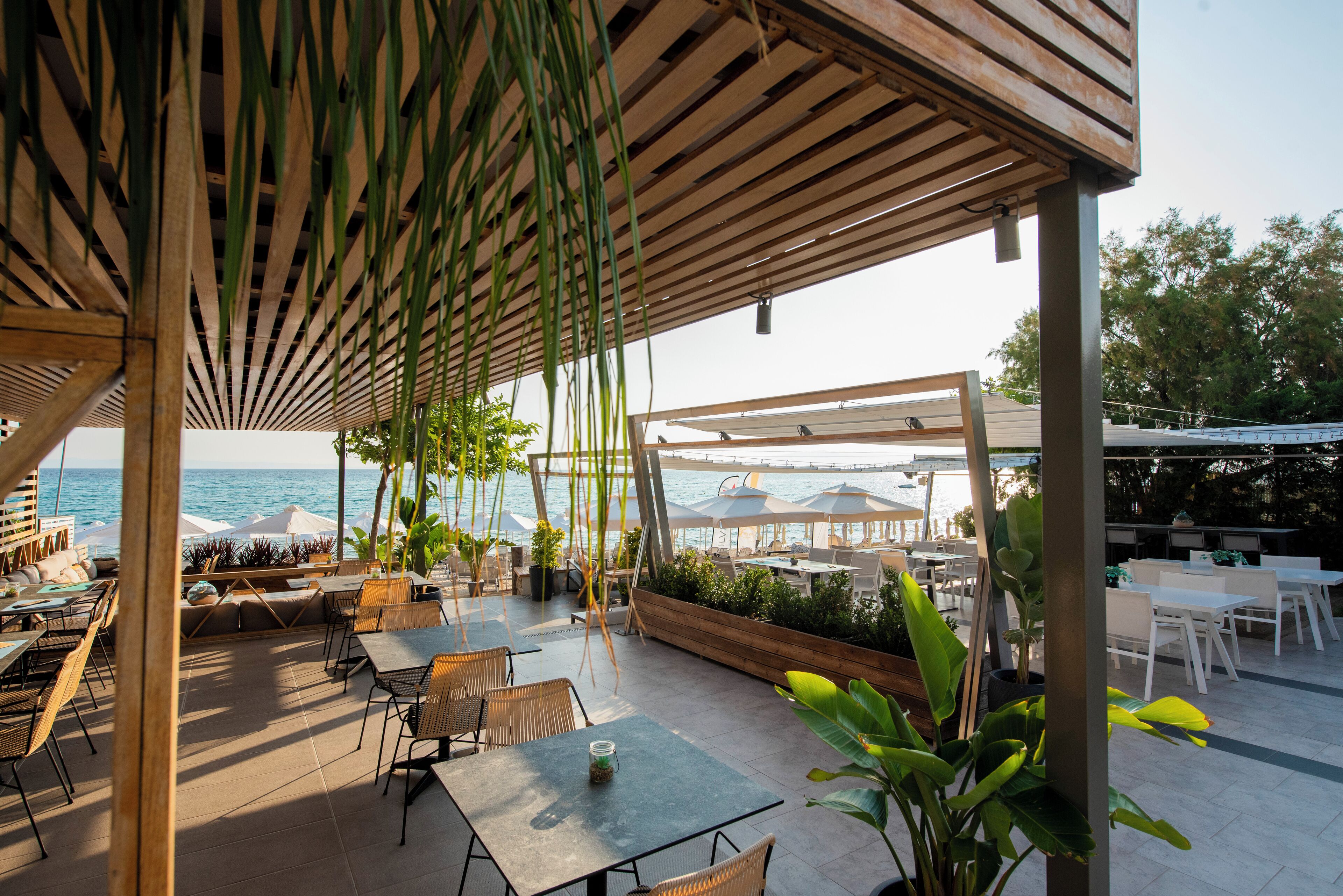 Un área de patio al aire libre con mesas y sillas, una pérgola de madera y vista al mar.