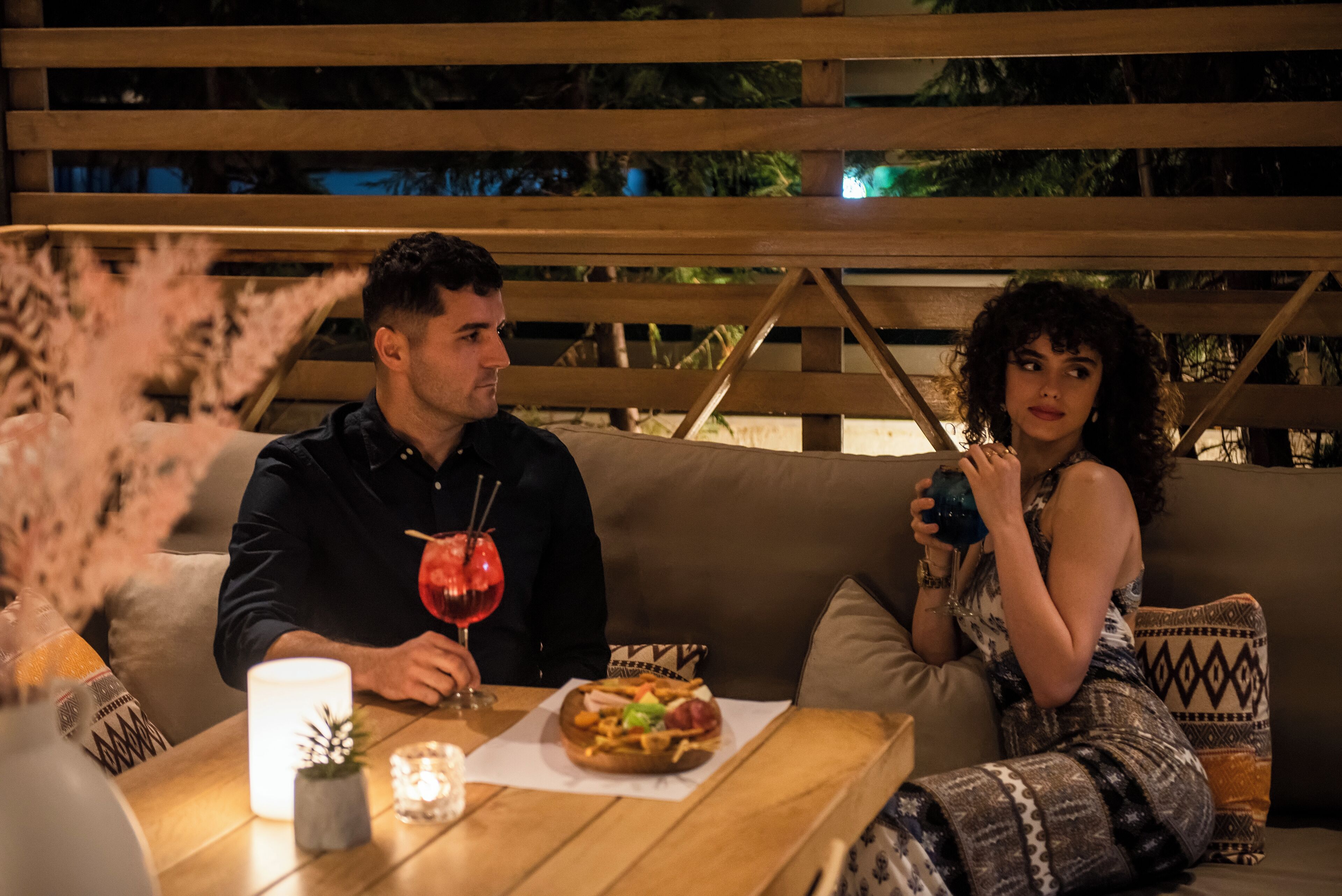 Una pareja sentada en una mesa al aire libre con comida y bebidas, rodeada de muebles de madera y cojines.