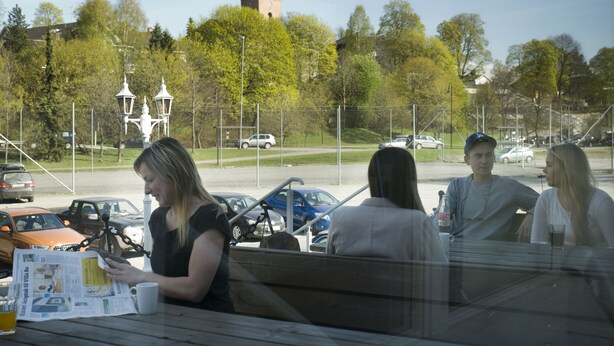 Un grupo de personas sentadas en una mesa al aire libre, con periódicos y bebidas, mirando hacia un estacionamiento con coches y una torre distante.