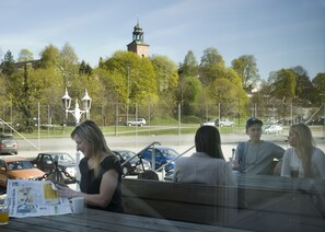 Eine Gruppe von Personen sitzt an einem Tisch im Freien, mit Zeitungen und Getränken, mit Blick auf einen Parkplatz mit Autos und einem entfernten Turm.