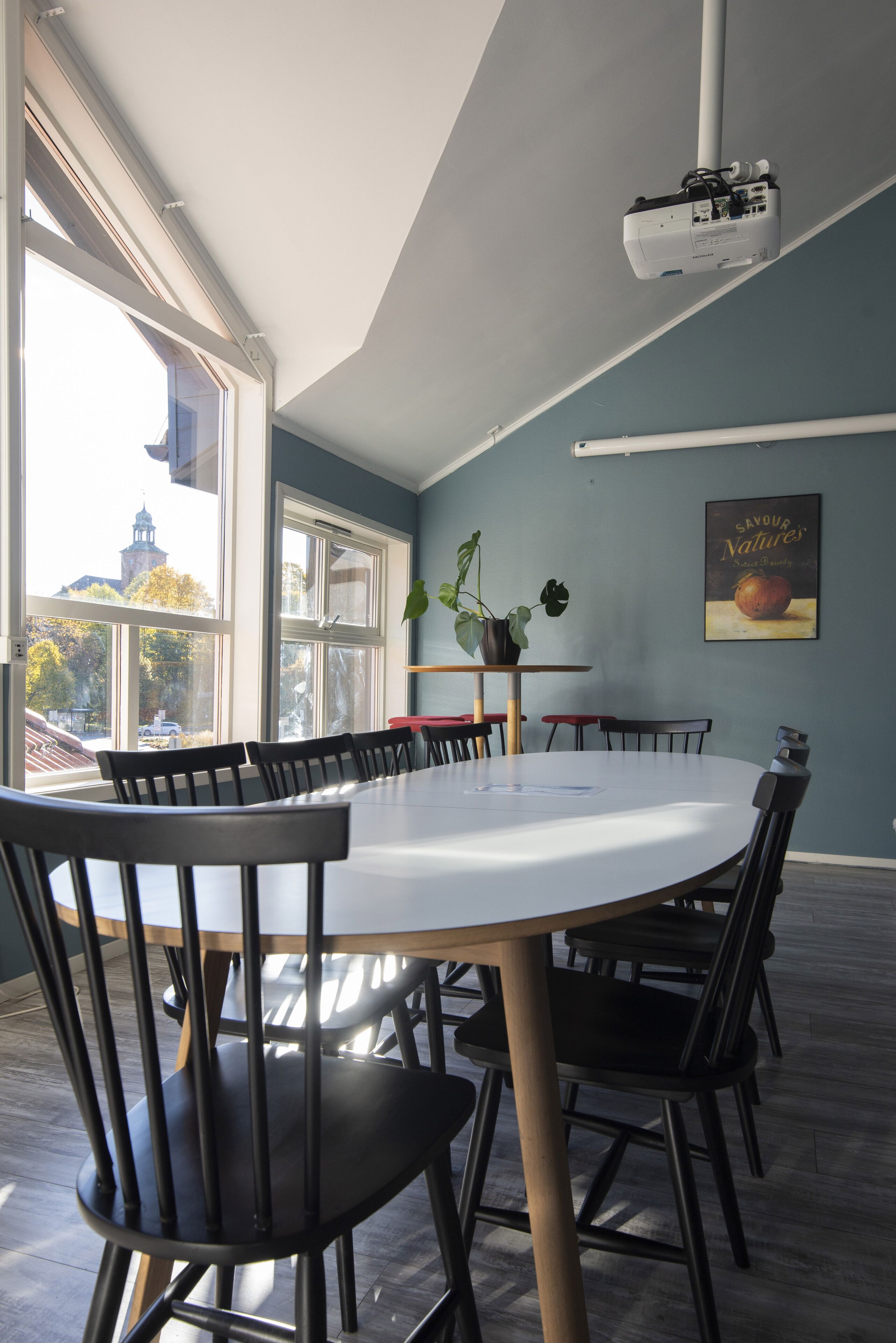 Een vergaderruimte met een grote tafel en stoelen, een projector die aan het plafond is bevestigd, en een raam met uitzicht op gebouwen en bomen.
