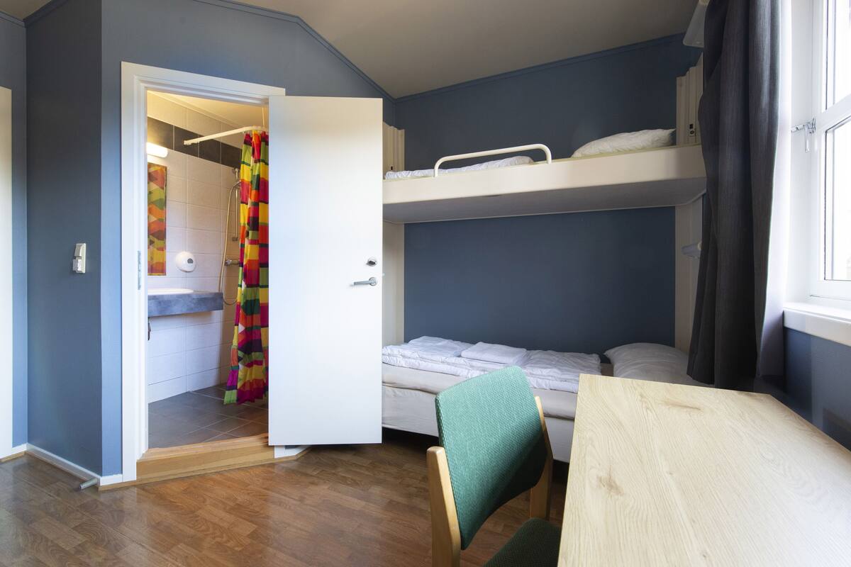Ein Zimmer mit Etagenbett, kleinem Tisch, Stuhl und Bad mit Duschvorhang, sowie einer Tür zu einem weiteren Raum.