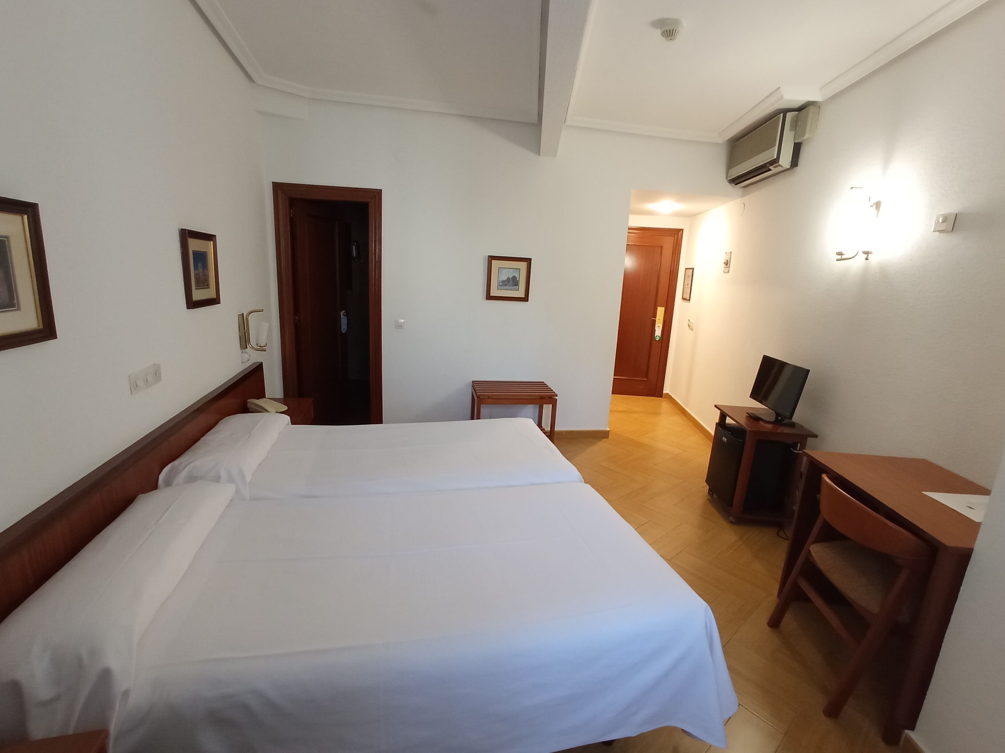 A hotel room with two beds, a desk, a chair, a TV, and a door leading to another room.