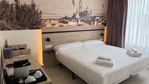 Et moderne hotelværelse med en stor seng, et sengebord med lampe, et lille skrivebord med kaffemaskine og et bybillede på væggen.