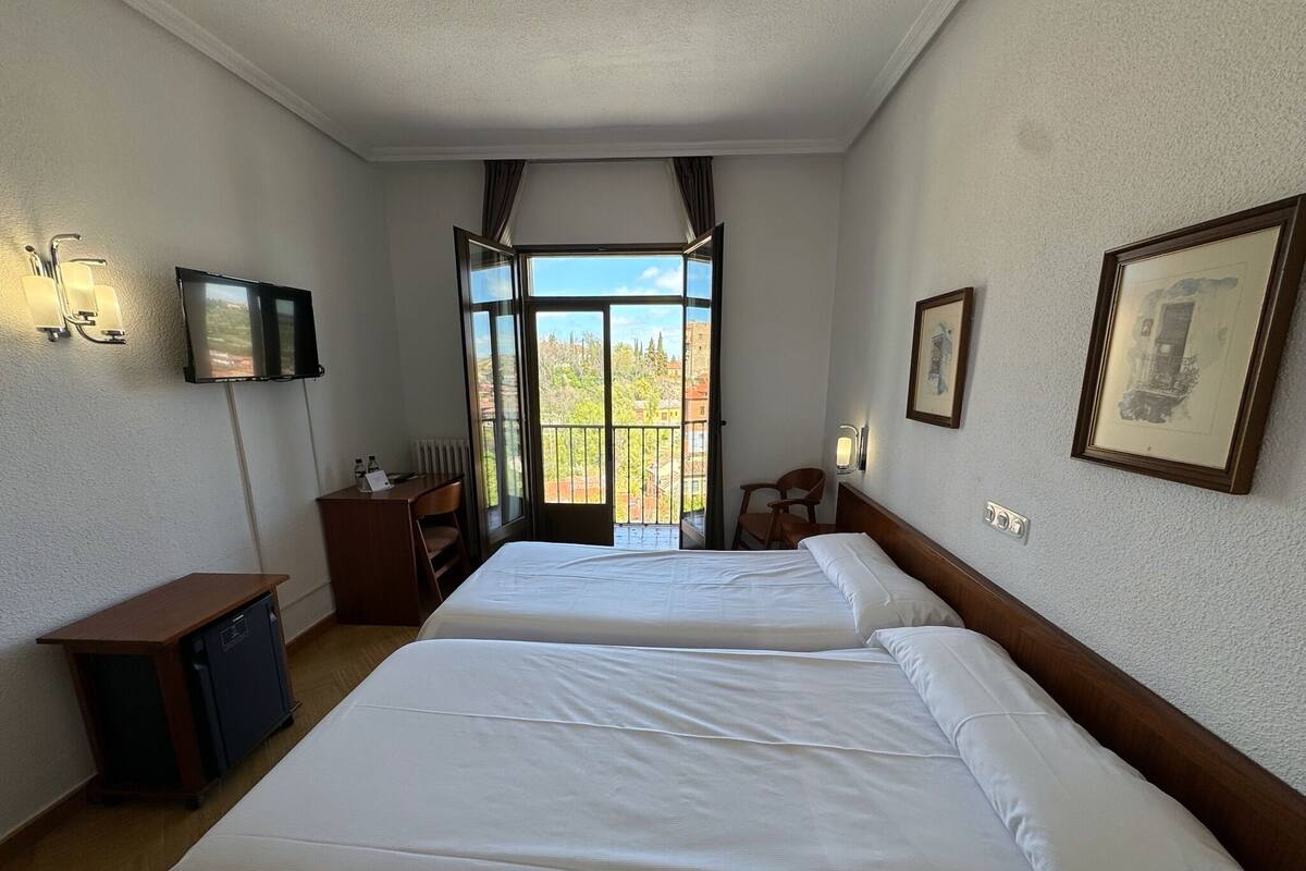 A hotel room with two beds, a desk, a chair, a TV, and a balcony with a view.