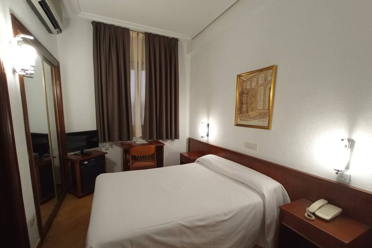 A hotel room with a bed, a desk, a chair, a television, and a painting on the wall.