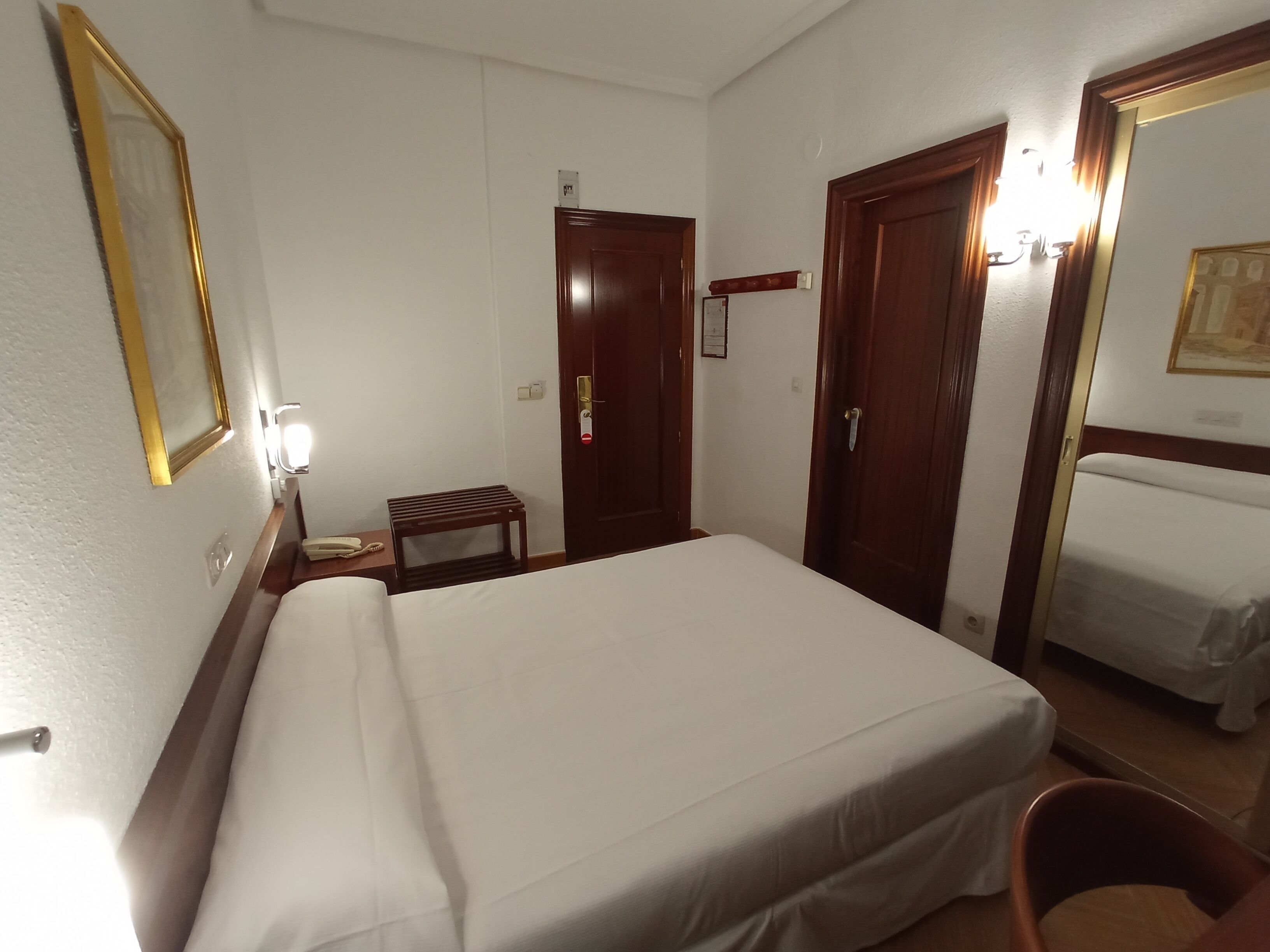 A hotel room with a large bed, a small bedside table with a lamp, a mirror, and a door with a handle.