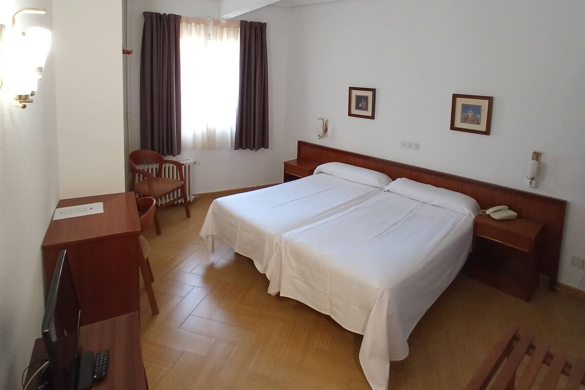 A hotel room with two beds, a desk with a computer, a chair, and a window with curtains.