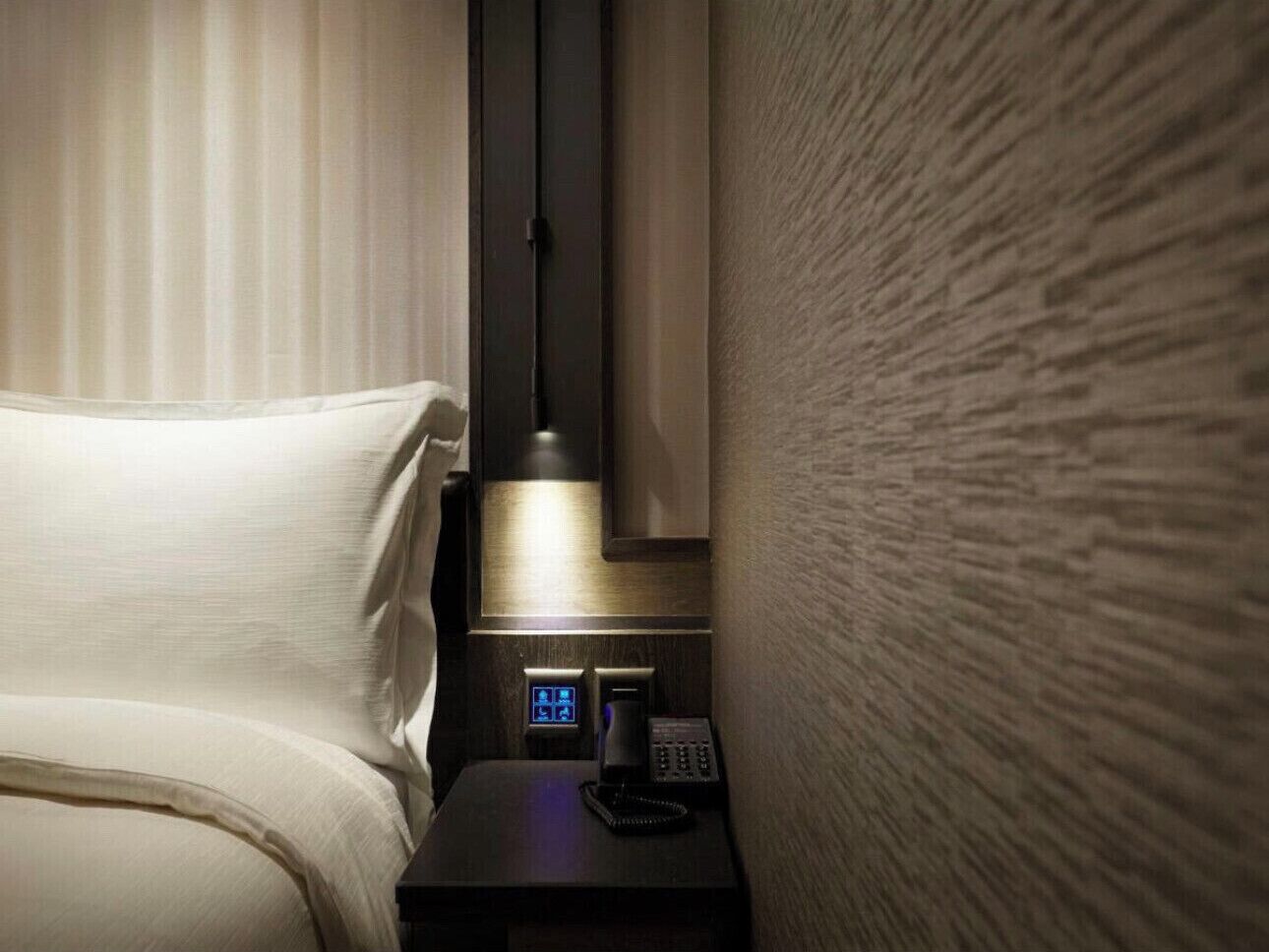 A hotel room with a bed, a bedside table with a lamp, a digital clock, and a telephone.
