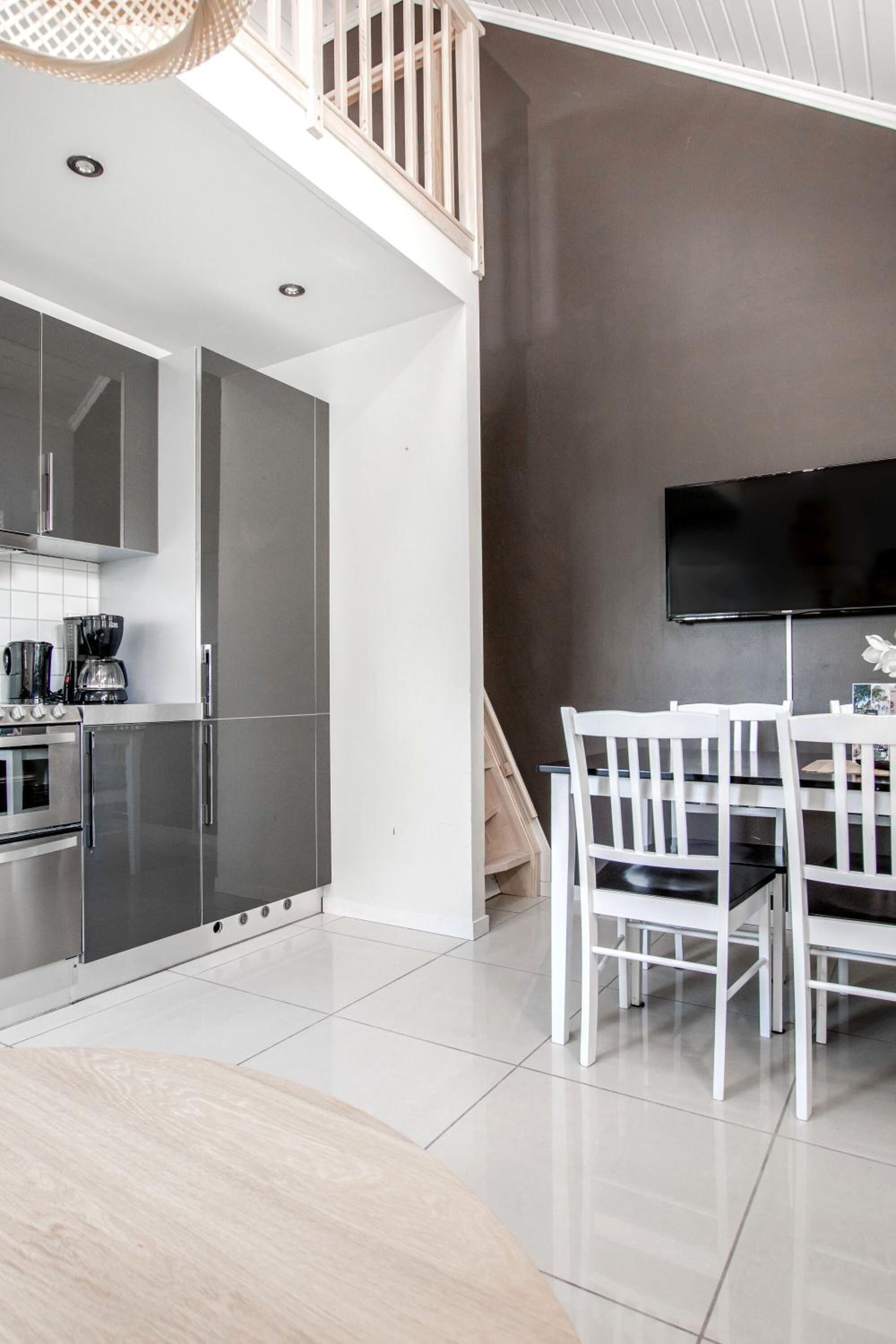 A modern kitchen with stainless steel appliances, a dining area with white chairs, and a large flat-screen TV mounted on the wall.