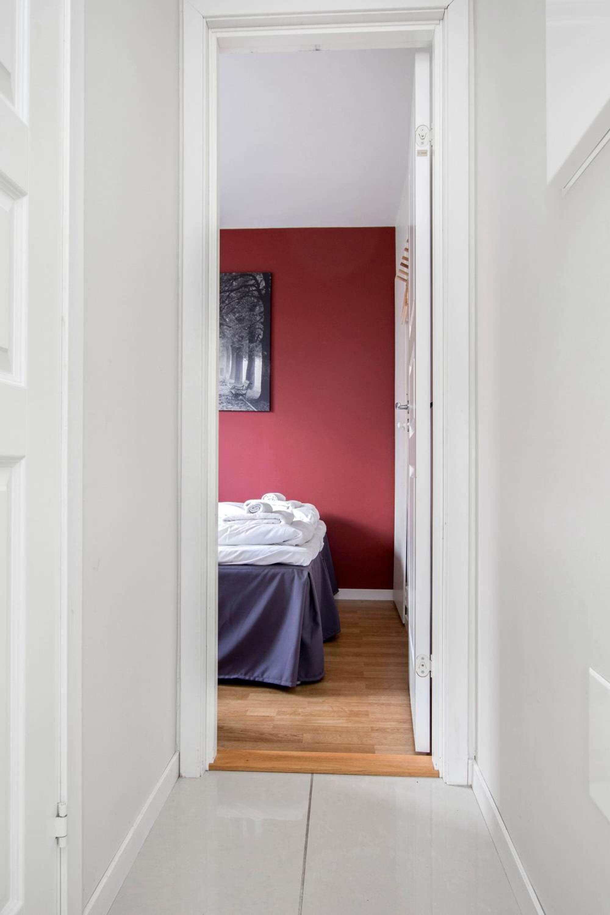 Un pasillo estrecho que conduce a un dormitorio con pared roja y cama con ropa blanca.