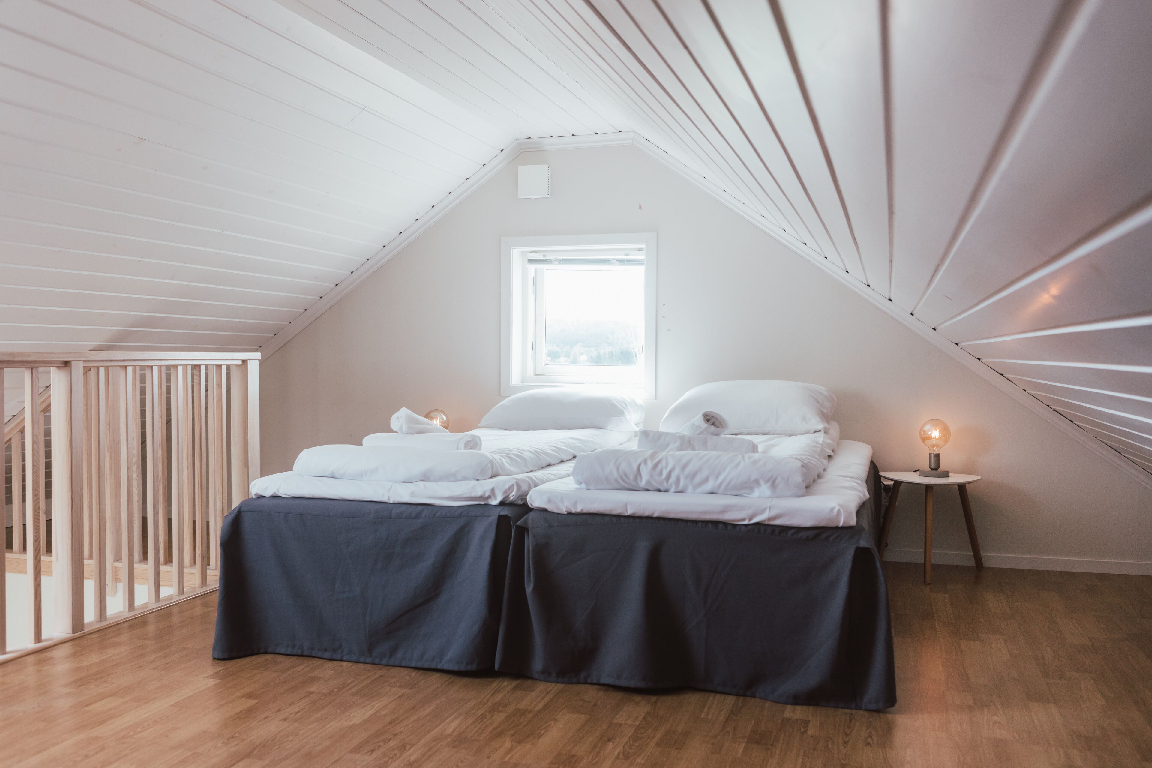 A bedroom with a sloped ceiling, a window, a bed with white linens, a bedside table with a lamp, and a wooden floor.