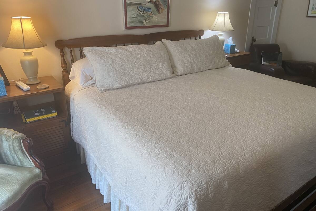 Un lit bien fait, recouvert d’une couette blanche, deux lampes de chevet, une table de chevet avec un livre et un téléphone, et une chaise avec un coussin.