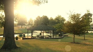 Un quiosco con mesas de picnic en un parque con árboles y un cuerpo de agua al fondo.