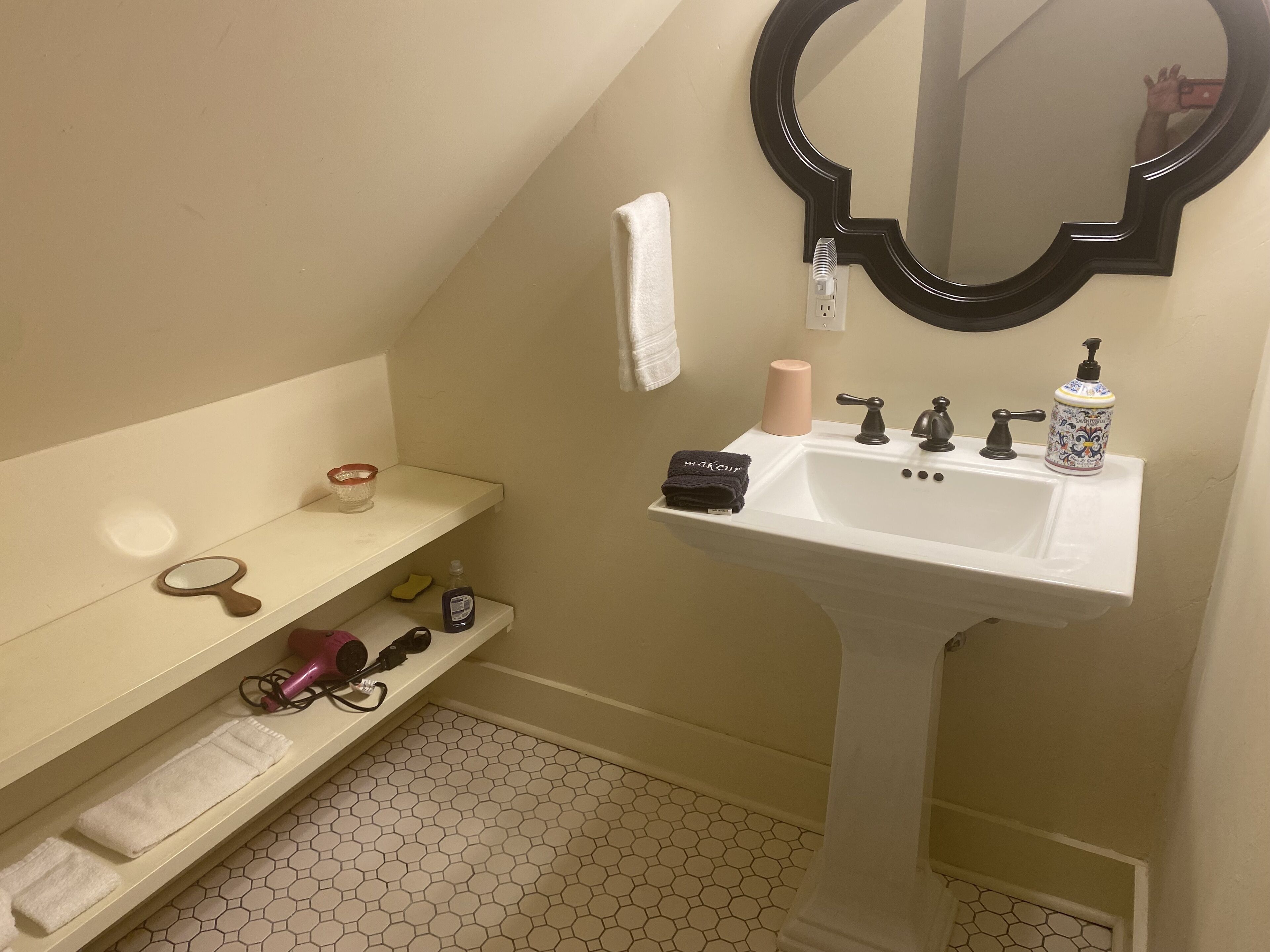 A bathroom with a white pedestal sink, a round mirror, a towel rack, and a shelf with toiletries.