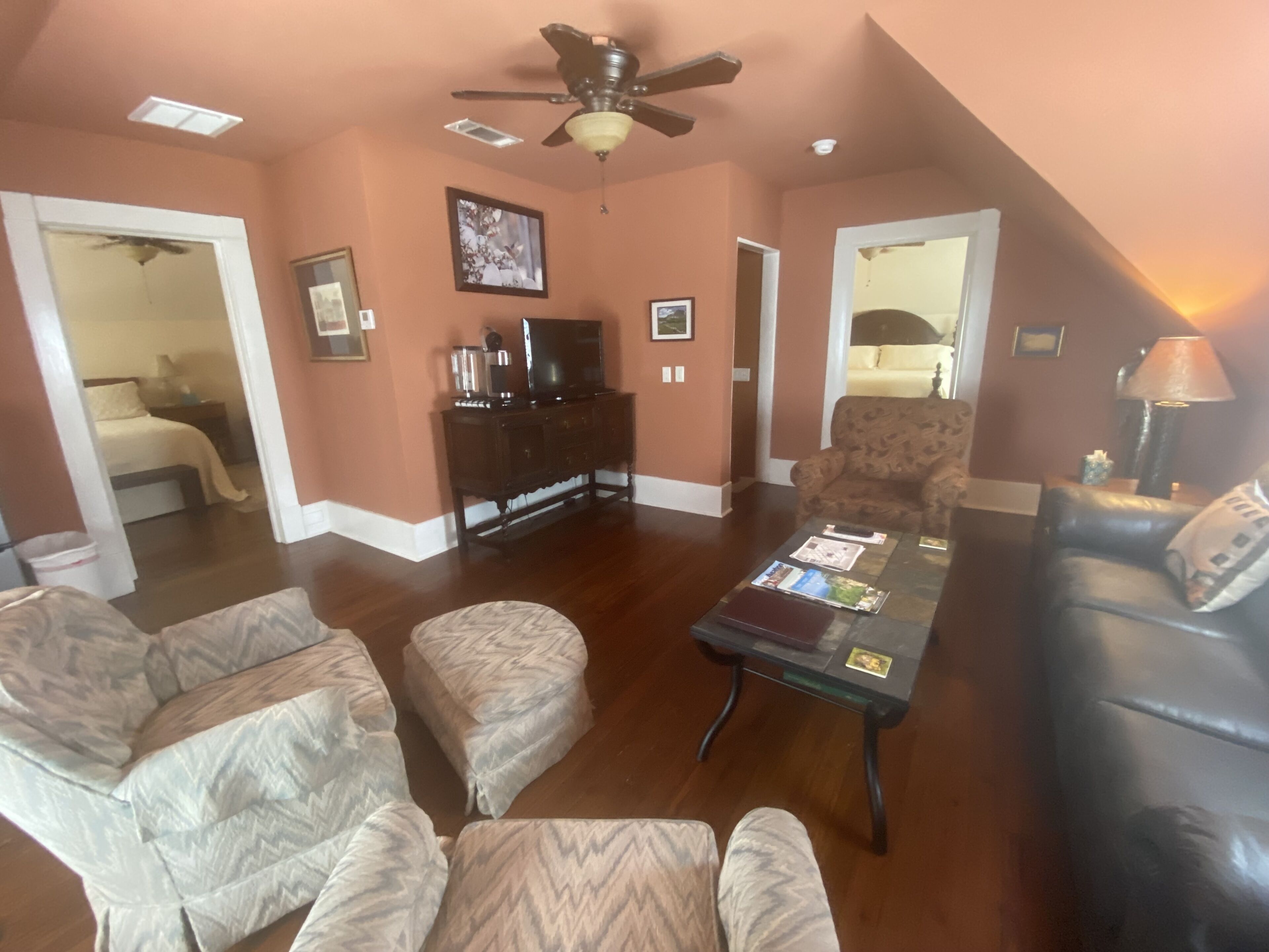 A spacious living room with a ceiling fan, a television, and a coffee table with magazines. There are two armchairs and a sofa.