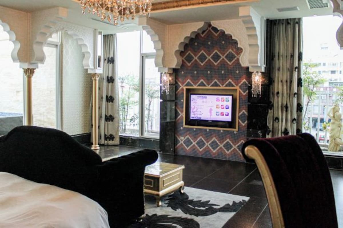 Habitación de hotel con un televisor grande montado en una pared decorativa, una cama con cabecera oscura y una ventana con vistas al exterior.