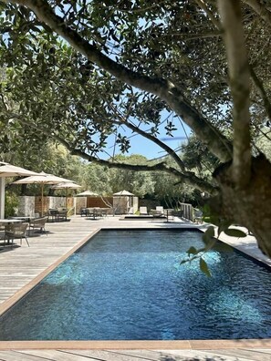 A rectangular swimming pool with a wooden deck surrounding it, outdoor seating, and trees in the background. - Hotel E Tre Stelle (Bonifacio)