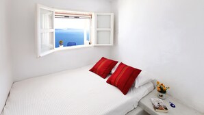 A bedroom with a bed, two red pillows, a nightstand with a book and a pen, a window with a view of the sea, and a ceiling light. - Menias Cave House (Santorini)