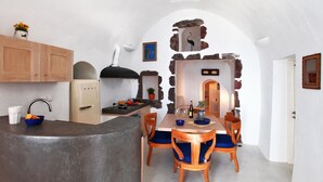 A compact kitchen with a central island, wooden cabinets, and a stone wall archway leading to a dining area with wooden chairs and a table. - Menias Cave House (Santorini)