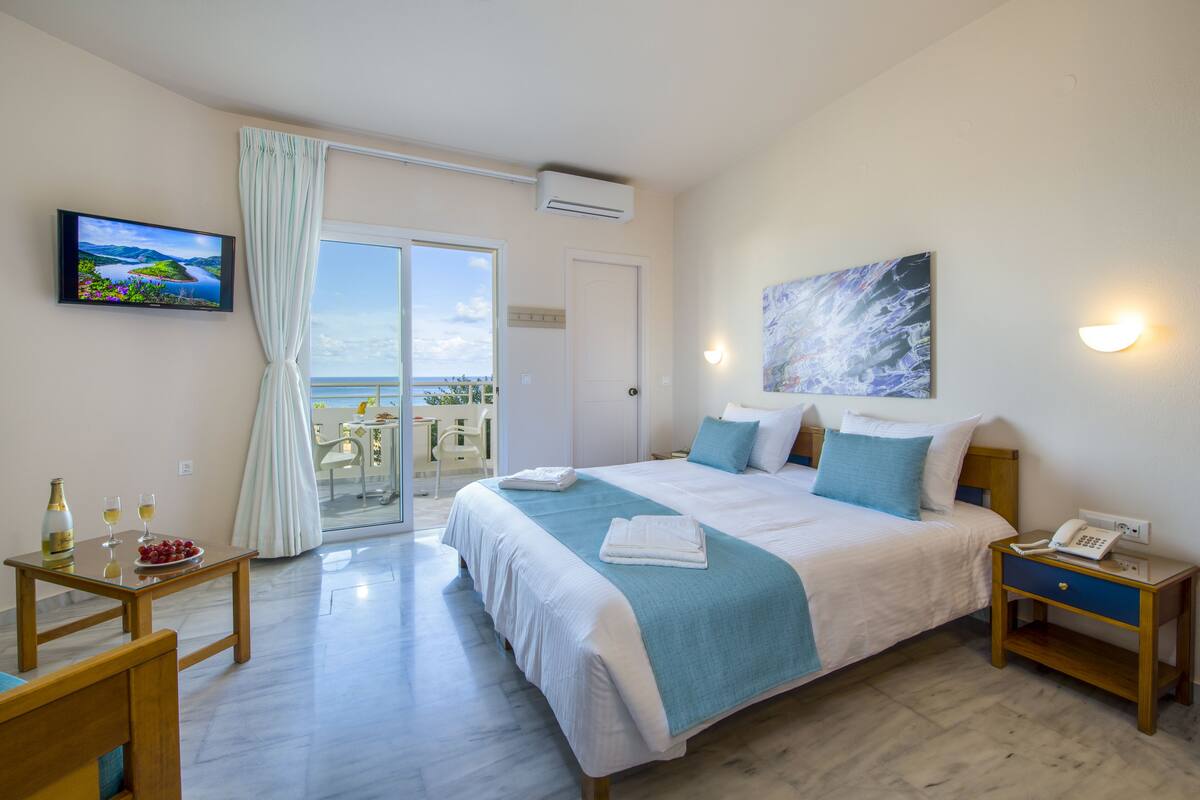Ein Hotelzimmer mit Bett, Nachttisch mit Telefon, Wandfernseher, einem Gemälde und Meerblick durch das Fenster.