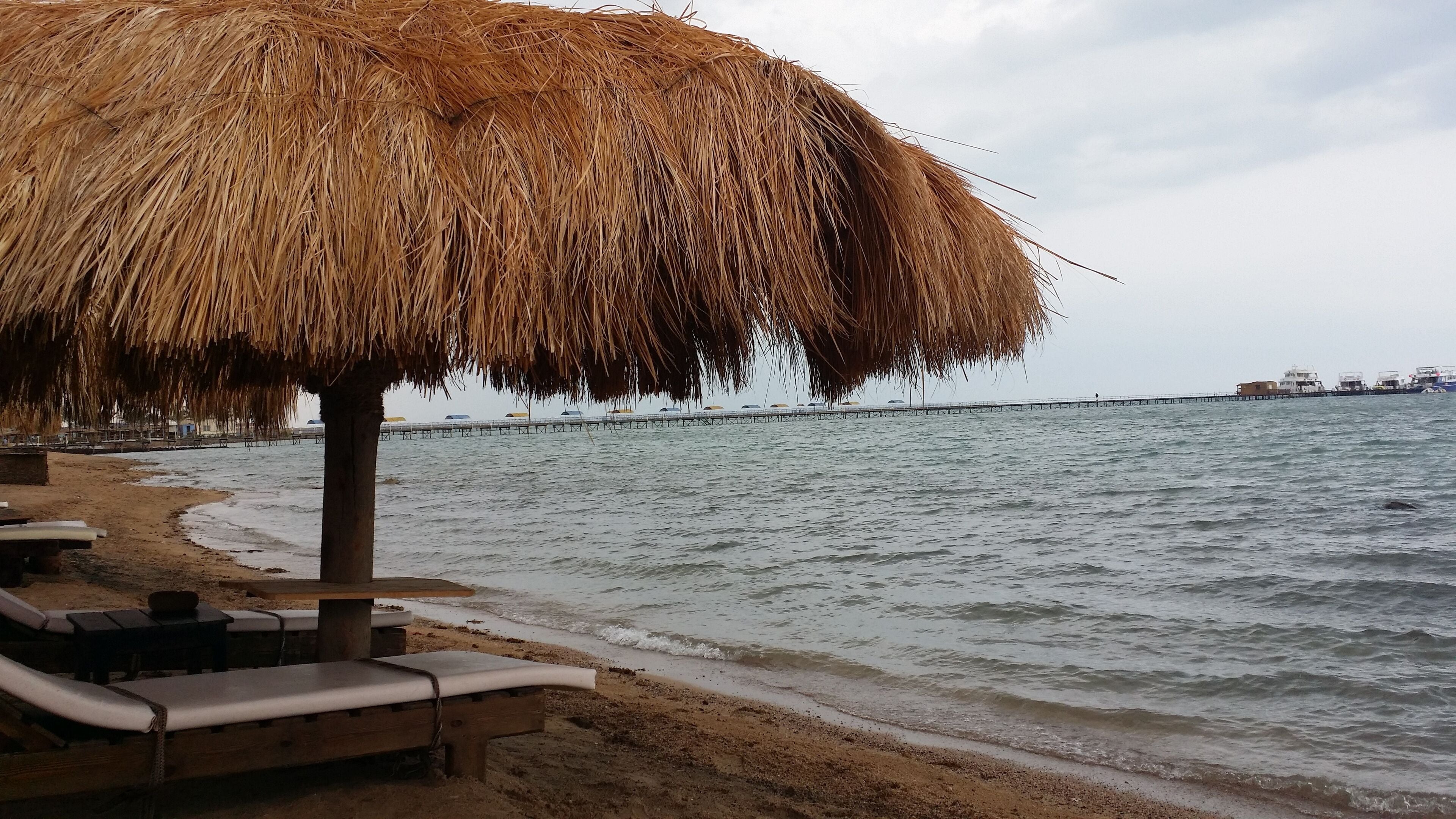 Une plage avec un parasol en chaume, des chaises longues en bois et une jetée en arrière-plan.