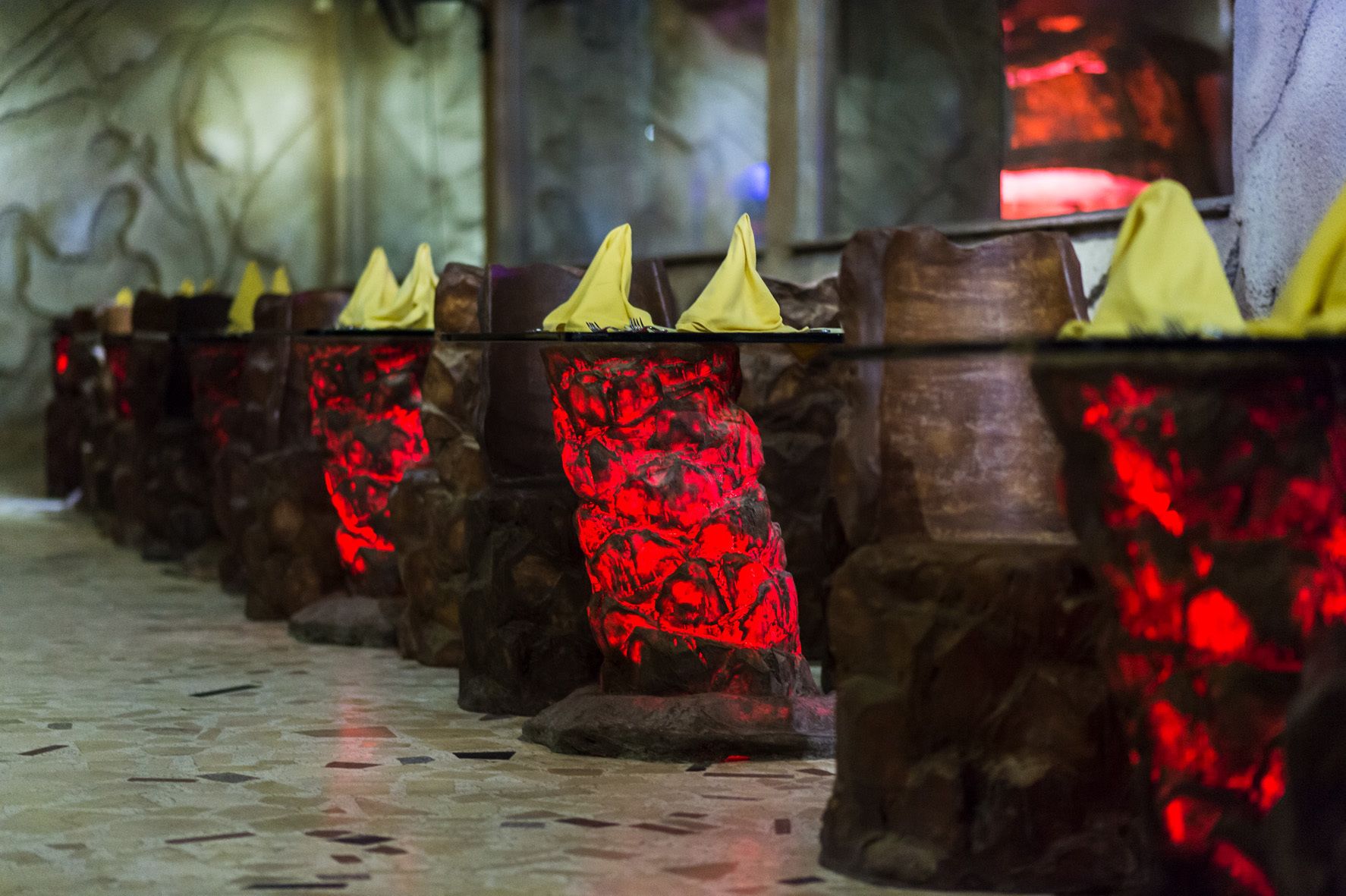 A row of stone tables with yellow napkins, set against a tiled floor and a textured wall.