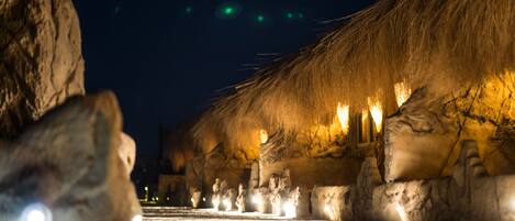 Fileira de construções com telhados de palha e janelas iluminadas, contra um céu escuro.
