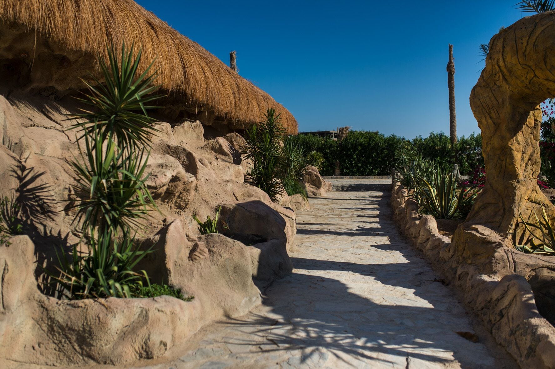 Un sentier bordé de rochers et de plantes, qui mène à une construction surmontée d’un toit de chaume.