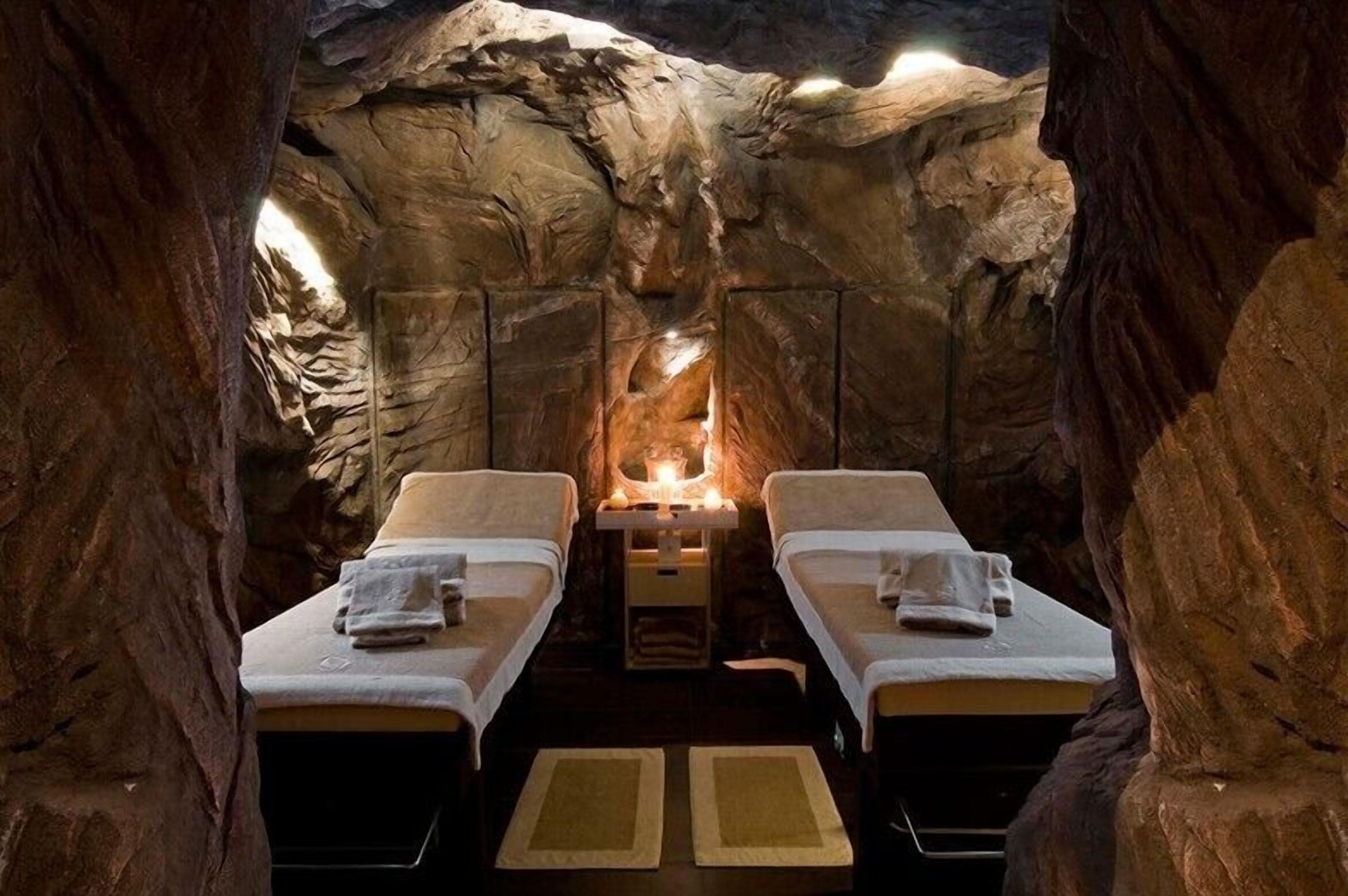Un cuarto de spa con dos camas, una mesa central con velas y una pared rocosa que recuerda a una cueva.